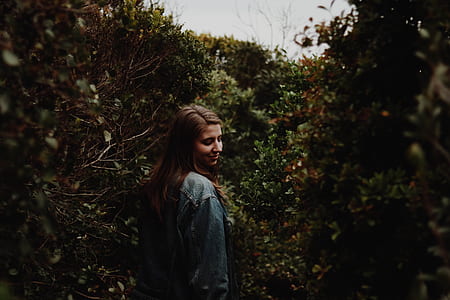 woman in blue long-sleeved top standing between green leaf plants thumbnail