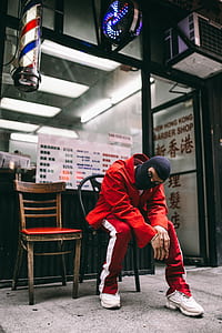 man wearing red jacket, pants, and white sneakers sitting on chair thumbnail