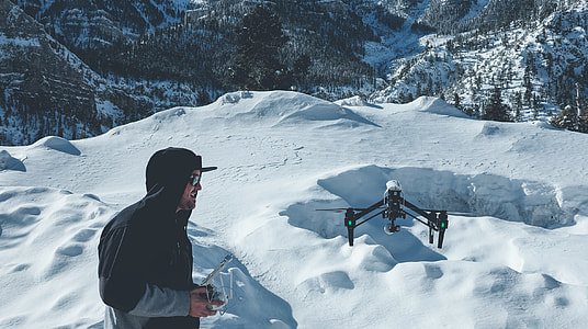 man wearing pull-over jacket infront of black quadcopter on snow field mountain thumbnail