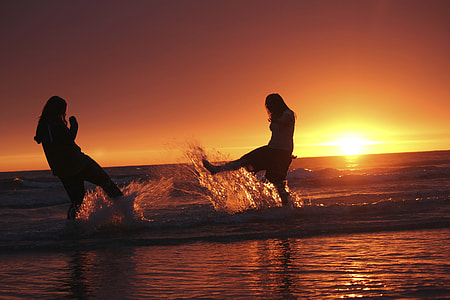 silhouette of two girls playing on sea shore kicking water during sunset thumbnail