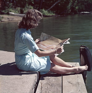 woman sitting on pavement while reading magazine beside body of water thumbnail