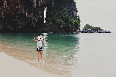 woman wearing white hat facing rock formation on body of water during daytime thumbnail