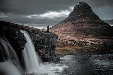 person standing atop waterfall near brown hill thumbnail