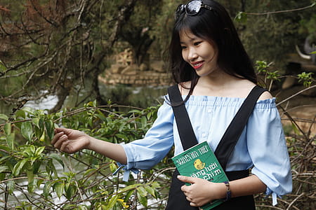 Woman Wearing Blue Off-shoulder Blouse Holding Book Next to Green Leaf Plant thumbnail