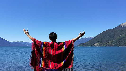 man wearing poncho with arms wide open in front of calm body of water and mountains during daytime thumbnail
