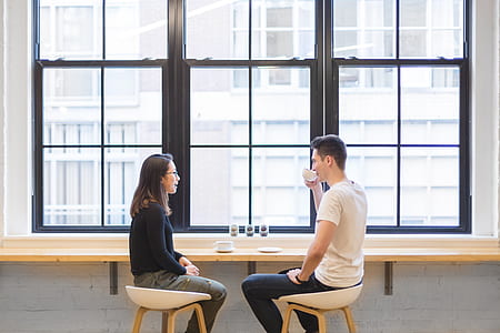 man wearing white t-shirt holding teacup on right hand talking to woman wearing black shirt while sitting on chairs thumbnail