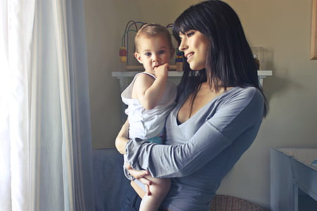 Woman Carrying Baby Boy Wearing White Tank Top Infront of White Curtain Inside the Room thumbnail