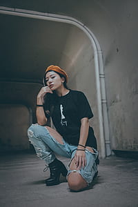 kneeling woman wearing black t-shirt, orange cap, black leather booties, and distressed blue denim jeans inside room with concrete walls thumbnail