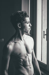 grayscale photo of topless man looking outside window thumbnail