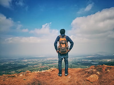 Man in Blue Dress Shirt and Blue Jeans and Orange Backpack Standing on Mountain Cliff Looking at Town Under Blue Sky and White Clouds thumbnail