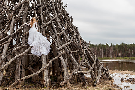 woman in white gown on gray pyramid formed tree branches beside lake thumbnail