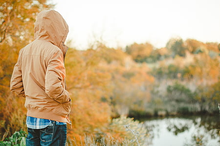 man in brown hooded jacket facing body of water during day thumbnail