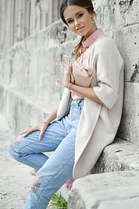 woman wearing beige cardigan and blue denim jeans leaning on gray concrete wall thumbnail