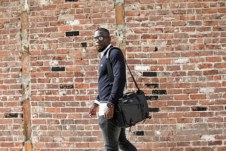 man wearing black shirt carrying bag standing beside brown wall thumbnail