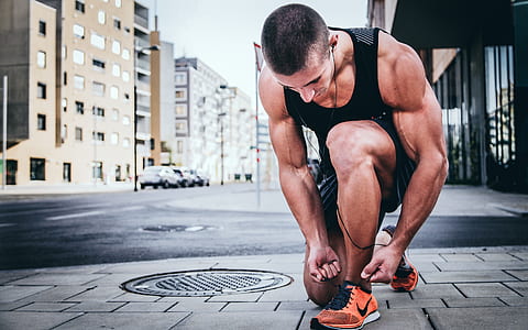 man wearing black tank top and black shorts tying his orange-and-black Nike running shoes beside manhole cover during daytime thumbnail