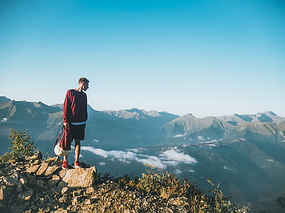 Man in Red Sweatshirt and Black Shorts Standing on Large Brown Rock on Top of a Mountain thumbnail