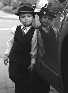grayscale photography of boy in dress shirt with vest and fedora hat leaning on car thumbnail