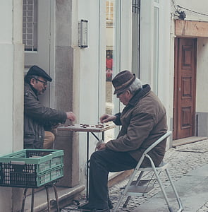 man sitting on step stool playing Chinese checkers with another man thumbnail