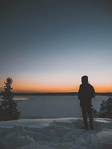 man standing on snow near tree overlooking body of water thumbnail