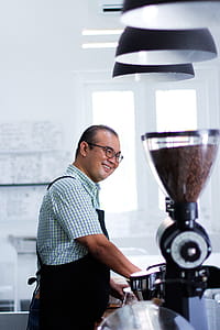 Man Wearing Plaid T-shirt And Black Apron in Front of Coffeemaker thumbnail
