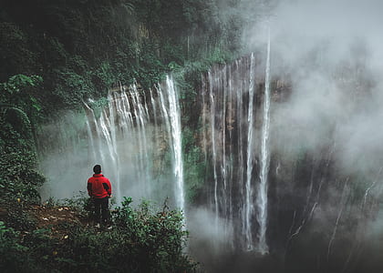 person wearing red jacket standing near mountain cliff facing on waterfall at daytime thumbnail