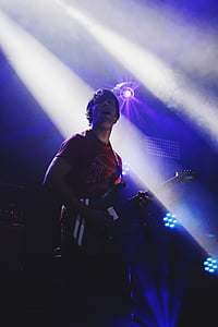 man wearing red crew-neck shirt playing electric guitar on stage with stage lights turned on thumbnail
