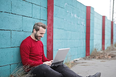 A young bearded man wearing a cherry red pullover sitting against a boundary wall smilingly looking at his laptop thumbnail