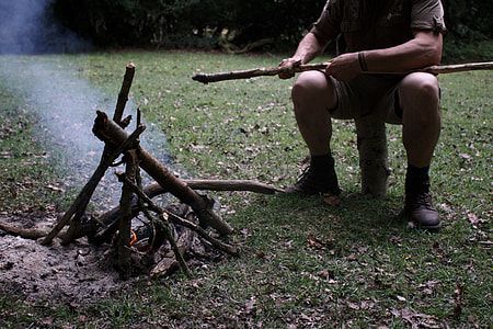 man sitting on log making fire with wood pile during daytime thumbnail
