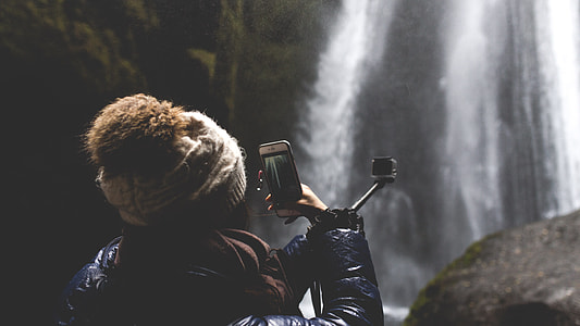 selective focus photography of woman holding smartphone while capturing waterfalls thumbnail