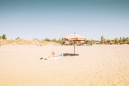 person lying on sand near pink patio umbrella during daytime thumbnail