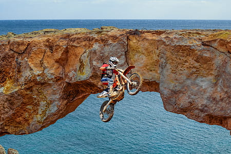 man riding motocross dirt bike in mid air near stone bridge and ocean during day thumbnail