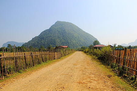 beige soil pavement between wooden fences with green mountain at distance under gray sky during day thumbnail