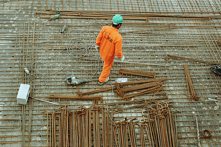 man in orange suit wearing teal hard helmet standing beside brown steel rods thumbnail