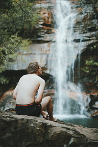 man sitting on black shorts sitting on rock facing waterfalls thumbnail