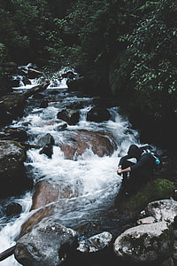 person sitting on rock beside body of water and trees during daytime thumbnail