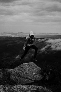 grayscale photo of a person jumping on rock on mountain landscape thumbnail