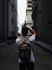 man wearing cap and backpack standing between buildings thumbnail