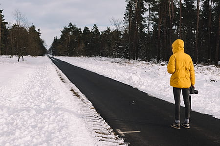 person wearing yellow hoodie standing on black concrete road near high trees under white sky during daytime thumbnail