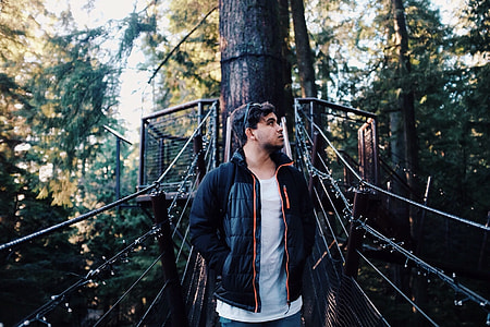 selective focus photography of man wearing black zip-up jacket and white top standing in brown bridge thumbnail
