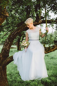 woman wearing white chiffon and lace short-sleeved a-line wedding dress on tree while holding white rose flowers bouquet thumbnail