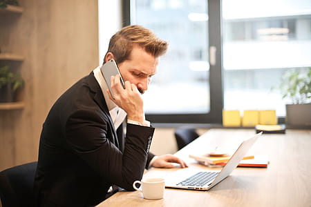 Young Adult Man talking on the cellphone and looking at his laptop while sitting at working place thumbnail