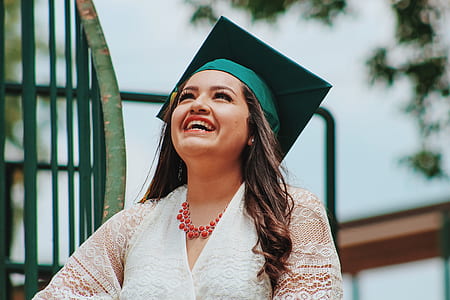 smiling woman wearing square green academic cap and white lace surplice top beside green metal spiral rail thumbnail