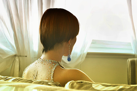 woman wearing gray studded lace sleeveless top and blue pendant hoop earrings sitting on sofa near white window curtain thumbnail