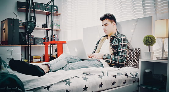 Man in Green, Blue, and Black Plaid Sports Shirt Sitting on Bed Using Silver Macbook thumbnail