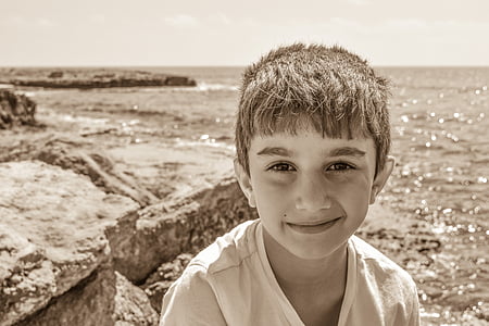sepia photography of boy standing on cliff mountain fronting body of water thumbnail