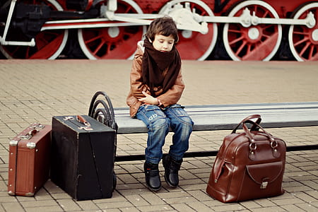 boy in brown jacket and blue jeans sitting on bench thumbnail