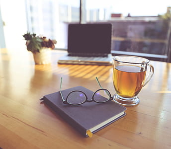 clear glass teacup filled with brown tea beside eyeglasses on top of brown wooden table thumbnail