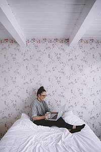 woman seats on white bed holding a white mug while using her laptop thumbnail