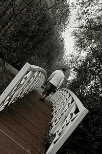 greyscale photography of man walking on bridge under trees thumbnail