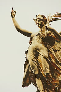 woman raising her right hand while holding branch with leaves on her left hand brass-colored statue low-angle photo thumbnail